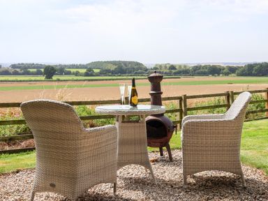 An outdoor seating area with two wicker chairs a glass table bottle and glasses overlooking fields at Bellwether in Helmsley