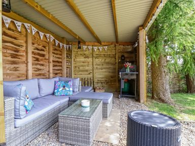 An outdoor covered seating area with a wicker sofa and glass table next to a barbecue grill at Bellwether in Helmsley