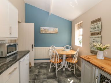 A kitchen dining area with a round wooden table and four chairs a microwave on a countertop and a wall decoration with cottage rules at Glasfryn in Llanfaethlu