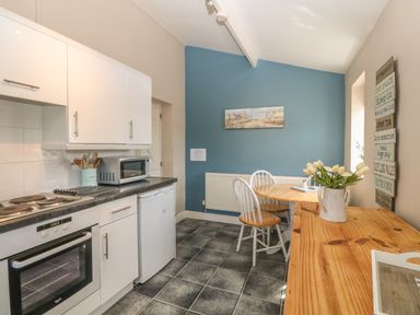 A kitchen with white cabinets a black countertop a microwave an oven a round wooden dining table with chairs a wooden sideboard and flowers in a jug on the sideboard at Glasfryn in Llanfaethlu