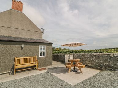 An outdoor patio area with a wooden bench against a house wall and a round wooden picnic table with an umbrella surrounded by a stone wall at Glasfryn in Llanfaethlu