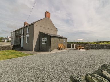 A grey stone and brick house with a gravel driveway bench and picnic table with umbrella at Glasfryn in Llanfaethlu