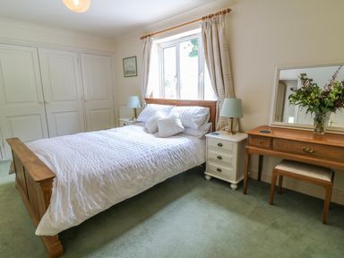 A bedroom with a wooden bed white bedding two bedside tables with lamps a wooden dressing table with a mirror and a vase of flowers at Wood View Cottage in Blackawton