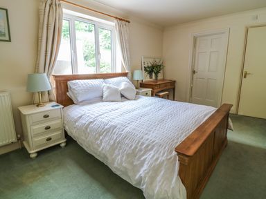 A bedroom with a wooden bed and white bedding a nightstand with a lamp a wooden desk with flowers and two doors at Wood View Cottage in Blackawton