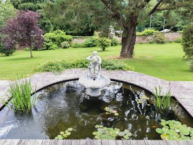 A garden with a circular pond featuring a statue fountain and lily pads surrounded by grass and trees at Wood View Cottage in Blackawton