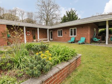 A garden with plants and flowers in front of a brick house with green lounge chairs and a picnic table at Wood View Cottage in Blackawton