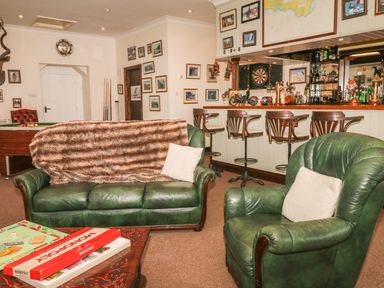 A living room with green leather sofa and armchair a coffee table with monopoly game a pool table and a bar with wooden stools at Wood View Cottage in Blackawton