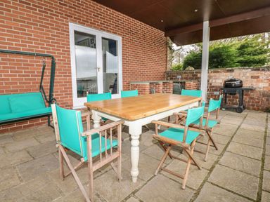 An outdoor patio area with a wooden table surrounded by turquoise chairs a barbecue grill and a swing seat at Wood View Cottage in Blackawton