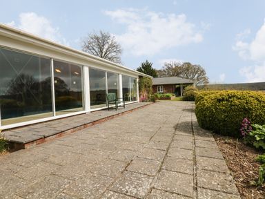 Patio area with stone tiles and a green bench next to a building with large glass windows at Wood View Cottage in Blackawton