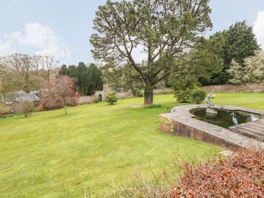 A garden with a large tree a stone pond with a statue and various bushes at Wood View Cottage in Blackawton