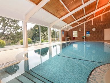 An indoor swimming pool with tiled floor windows overlooking outdoor trees and bushes at Wood View Cottage in Blackawton