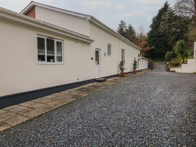A gravel driveway alongside a single story building with potted plants near the doors and garden area at Wood View Cottage in Blackawton