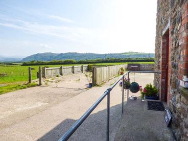 An outdoor patio area with hanging plants a potted bush and a metal railing next to a stone building at Muncaster View in Carleton near Ravenglass