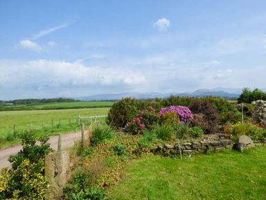 A garden with flowering plants and shrubs near a dirt path with fields and hills in the background at Muncaster View in Carleton near Ravenglass