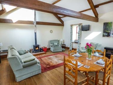 A living room with wooden beams a wood stove two armchairs a sofa a dining table with chairs and flowers at Muncaster View in Carleton near Ravenglass