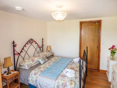 A bedroom with a double bed with floral bedding and blue runner beside a wooden door with a lamp on a wicker table and a vase of flowers on a dresser at Muncaster View in Carleton near Ravenglass