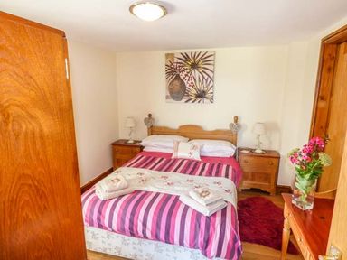 A bedroom with a double bed striped pink bedding bedside tables with lamps a wooden wardrobe and a wooden dressing table with a vase of flowers at Muncaster View in Carleton near Ravenglass
