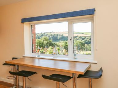 A dining area with a wooden table and three black bar stools by a window with a view of greenery at Rose Cottage in Barnstaple