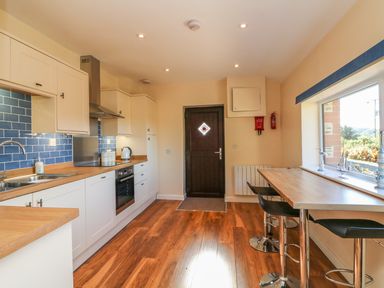 A kitchen with white cabinets blue tile backsplash wood countertops a wooden door and a wooden breakfast bar with stools at Rose Cottage in Barnstaple
