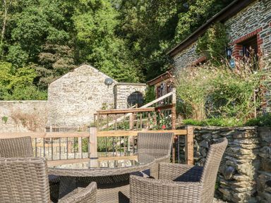 An outdoor patio area with wicker chairs and a glass table in front of a stone building with plants at Rose Cottage in Barnstaple