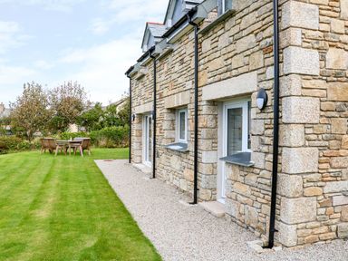 A stone house exterior with a gravel path and green lawn with outdoor wooden table and chairs at Myles View in Pendeen