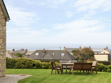 A garden area with wooden table and chairs overlooking rooftops and sea at Myles View in Pendeen