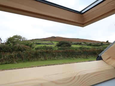 A view of a hillside with grass and bushes seen through an open wooden roof window at Myles View in Pendeen