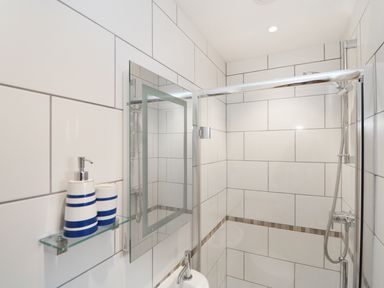 A bathroom with white tiled walls a glass shelf with two striped dispensers a mirror and a shower at Myles View in Pendeen