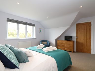A bedroom with a bed covered with a teal blanket and pillows a blue chair a wooden dresser with a television a window and wooden door at Myles View in Pendeen