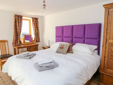 A bedroom with a bed with white bedding and a purple headboard a wooden dressing table with a mirror and plaid curtains at Apple Tree Cottage Bleatarn near Kirkby Stephen