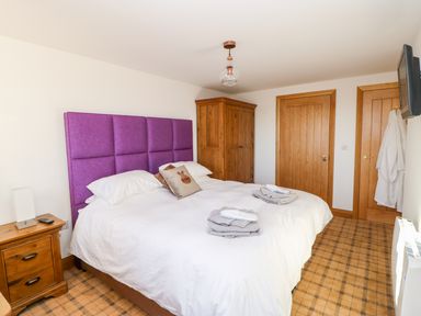 A bedroom with a bed with a purple headboard two wooden doors a wooden wardrobe a nightstand a wall-mounted television and folded towels on the bed at Apple Tree Cottage in Bleatarn near Kirkby Stephen