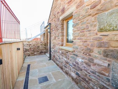 A stone walkway with a window on a stone wall and a red metal railing next to a wooden fence at Apple Tree Cottage in Bleatarn near Kirkby Stephen