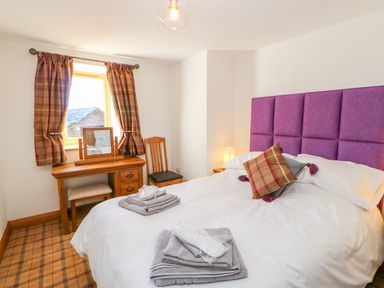 A bedroom with a bed with purple headboard and folded towels on it a wooden desk with a chair and a window with plaid curtains at Apple Tree Cottage in Bleatarn near Kirkby Stephen