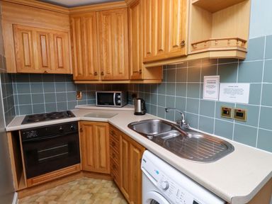 A small kitchen with wooden cabinets a black oven and stovetop a microwave kettle sink and washing machine at Bracken Lodge in Wooler