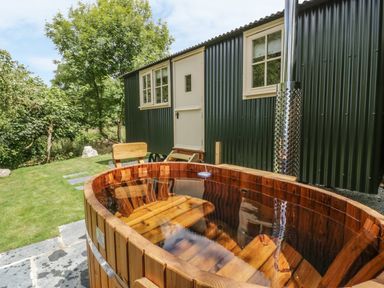 An outdoor wooden hot tub with bench seating next to a green corrugated metal building with white windows and door at Orchard Burlawn near Wadebridge