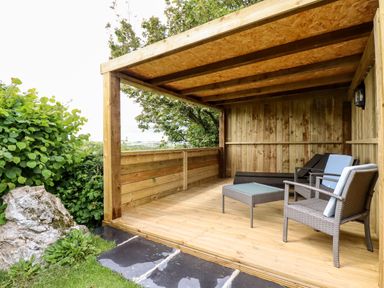 A wooden outdoor covered seating area with chairs a table and greenery at Orchard Burlawn near Wadebridge