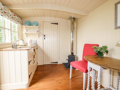 A small kitchen area with a wooden floor a red chair a round table a wood stove and shelves with plates at Orchard Burlawn near Wadebridge