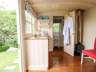 A small kitchen area with a sink kettle shelves and a view of a bathroom with a bathrobe on the door and a red chair beside a wood stove at Orchard Burlawn near Wadebridge