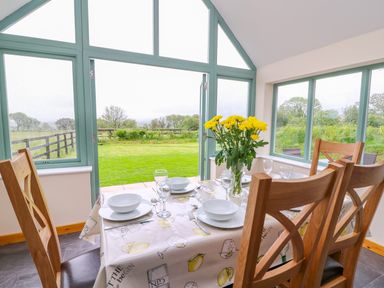 A dining table set with white bowls glasses and yellow flowers by large windows overlooking a garden at Bwthyn Eisteddfa Fach in Brynberian near Newport