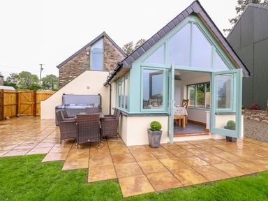 An outdoor patio area with a glass dining table and wicker chairs next to a small stone and cream building with open glass double doors at Bwthyn Eisteddfa Fach in Brynberian near Newport