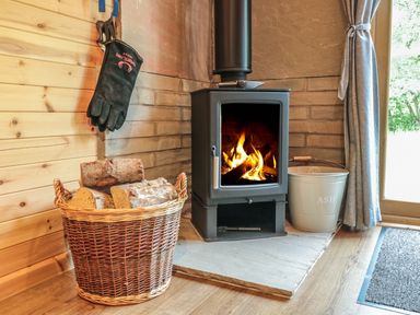 A wood burning stove with fire inside next to a basket of firewood and an ash bucket by a window at Dingle Den in Craswall