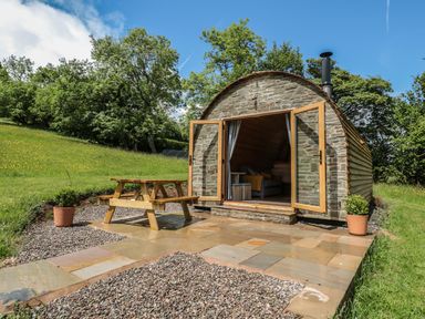 A stone cabin with open wooden doors a wooden picnic table on a paved patio surrounded by grass and trees at Dingle Den in Craswall