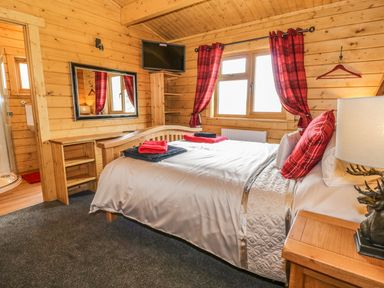 A bedroom with a bed cushions and towels wooden walls a window with red curtains and a mounted television at Manor Farm Lodges - Red Kite Lodge in Newtown