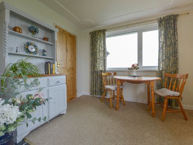 A dining area with a wooden table and two chairs next to a window with curtains and a cabinet with shelves and plants at The Quillet in Pendeen
