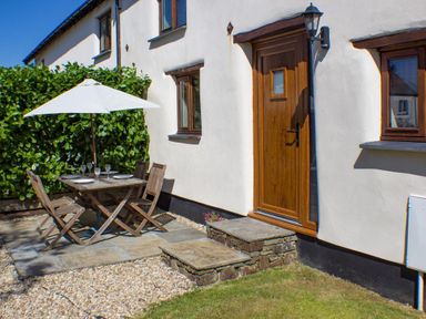 An outdoor patio area with a wooden table and chairs under a white umbrella next to a wooden door and windows at Post House in Holsworthy
