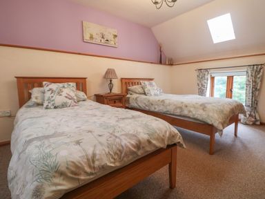 A bedroom with two single beds a wooden nightstand with a lamp a skylight and a window with curtains at Post House in Holsworthy