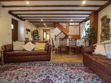 A living room with two brown leather sofas a dining table with chairs and floral tablecloth wooden beams on ceiling and a patterned rug on floor at The Croft in Holsworthy