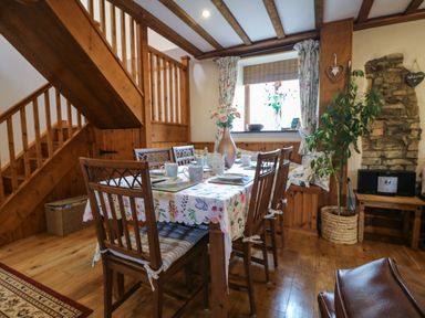 A dining area with a wooden table and chairs set with plates and mugs next to a wooden staircase and window at The Croft in Holsworthy