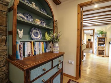 A wooden cabinet with books and decorative plates next to a doorway leading to a room with wooden furniture and a plant at The Croft in Holsworthy