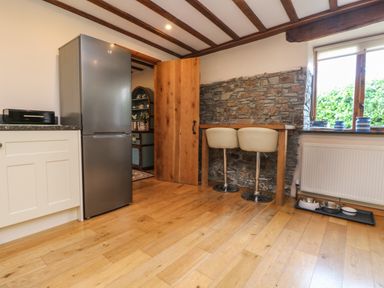 A kitchen area with a stainless steel refrigerator white cabinets wooden floor a stone wall a wooden table with two white chairs a window with blue jars and pet bowls on the floor at The Croft in Holsworthy